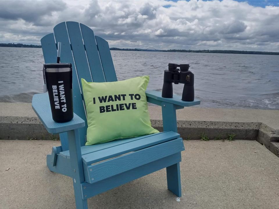 A blue Adirondack chair next to a lake holds a pair of binoculars and a pillow that says "I want to believe."