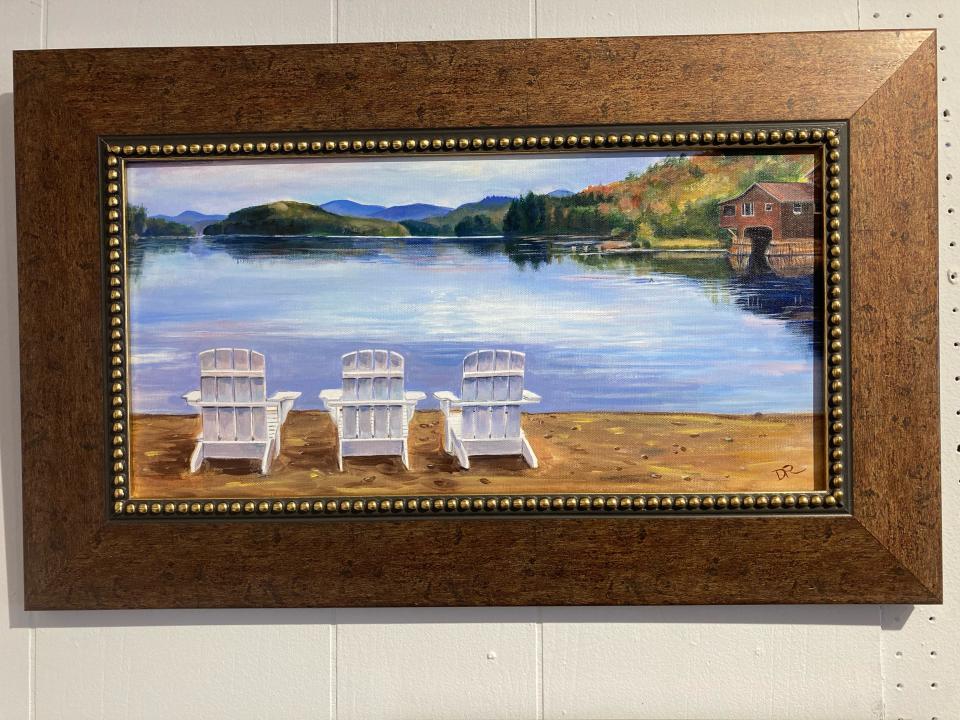 A lovely painting of Adirondack Chairs next to a body of water with a brown boat house and rolling hills along the shore.