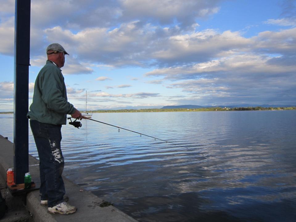 A man fishes from a pier while looking at mountains in the distance.