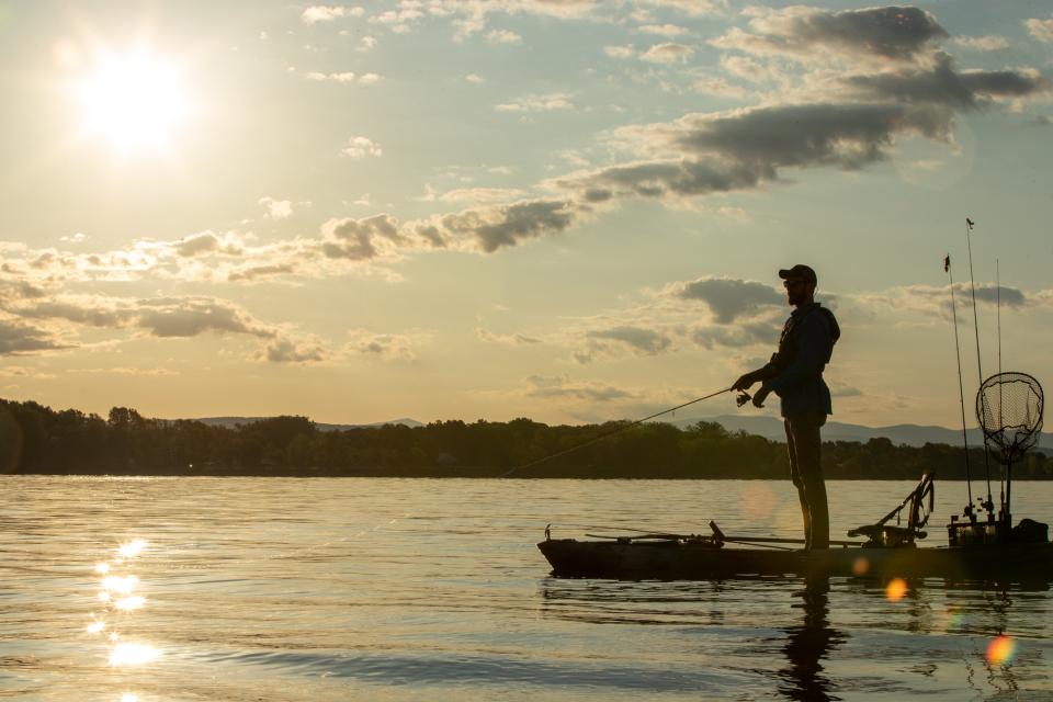 A man fishes from his boat on the water at dusk with mountains in the background.