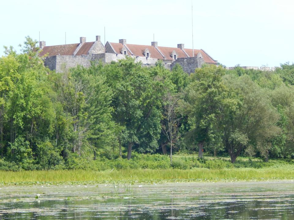 An old stone fort with brick-red roof rises above trees and a marshy shoreline.