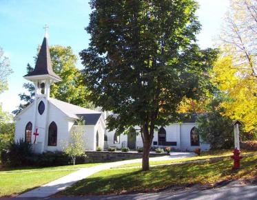 A white church with grey steeple and a large pine tree out front.