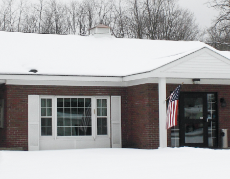 A squat brick building with white trim and covered entry way with large lawn blanketed in snow.