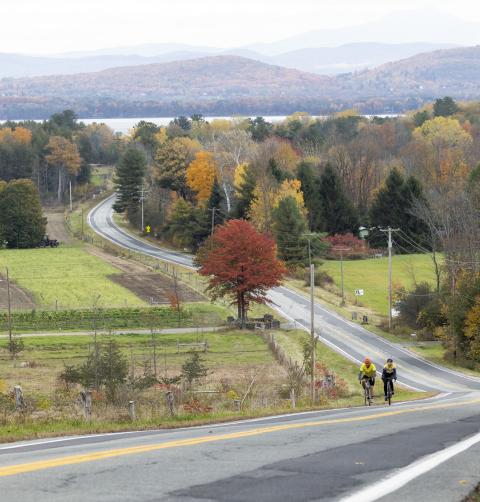 A view of a winding road in autumn, with brightly colored foliage and Lake Champlain in the distance