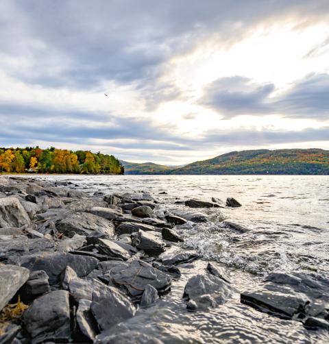 A view of Lake Champlain, with a rocky shoreline in the foreground and brilliantly colored fall foliage on the distant shore