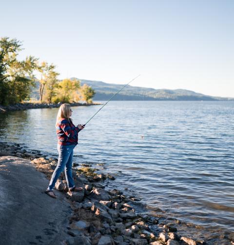 A woman casts a line off of a rocky shore of a lake. 