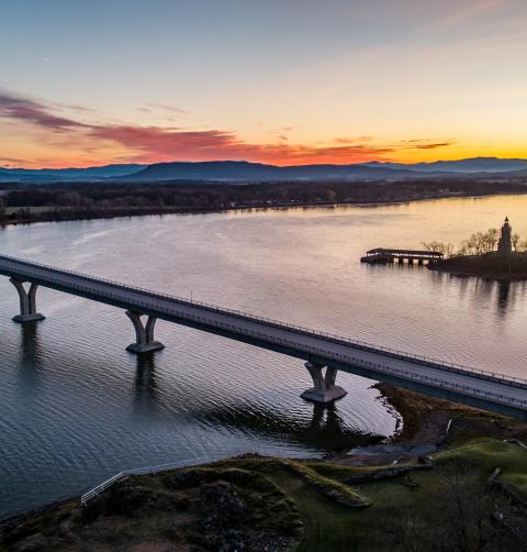An aerial view of a bridge spanning a narrow lake, with sunset on the horizon.