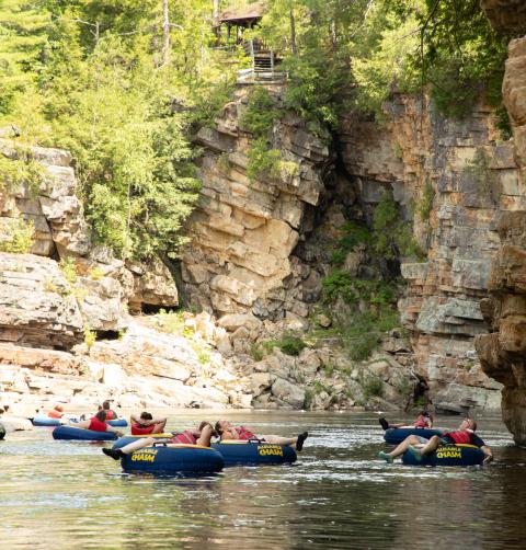 A group of people float on tubes in the river between a rocky chasm. 