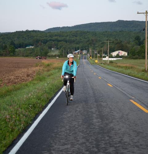 A woman wearing a blue jacket rides a bike toward the camera along a road in the Lake Champlain Region