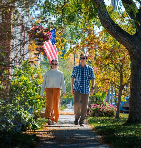 A man and woman walk on a neighborhood road covered in fall foliage.