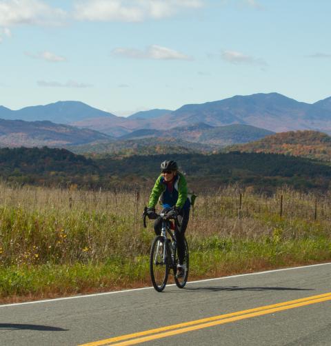 A woman rides her bike on a road with mountains in the background.