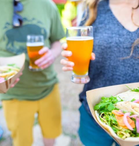A man and woman walk with tacos and beer.