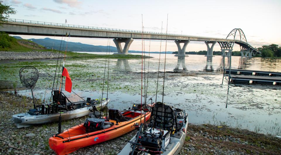 Kayaks fitted with trolling rods lined up on the edge of the water by a bridge.