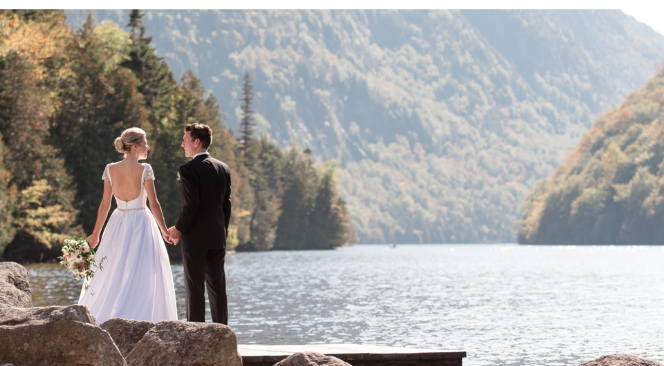 A bride and groom stand on a dock by the water surrounded by pine covered mountains.
