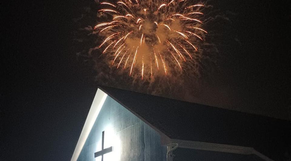 An illuminated cross on the side of a building with a firework exploding above.