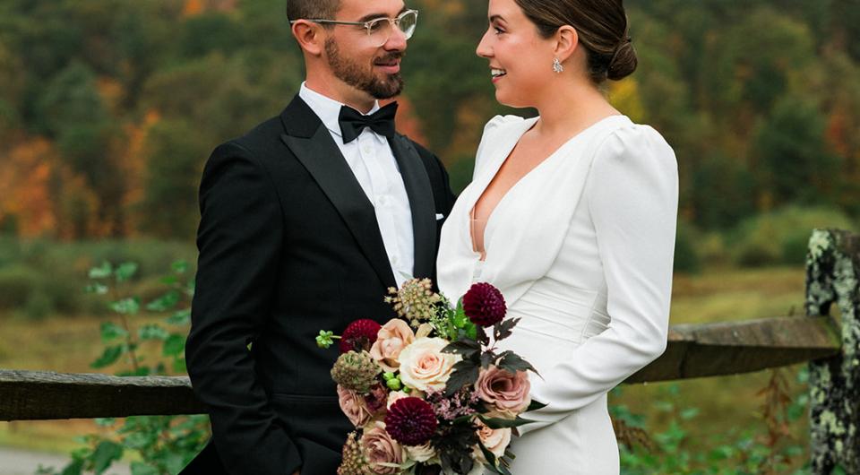 Bride and groom stand near a fence while she holds a bouquet of deep colored roses.