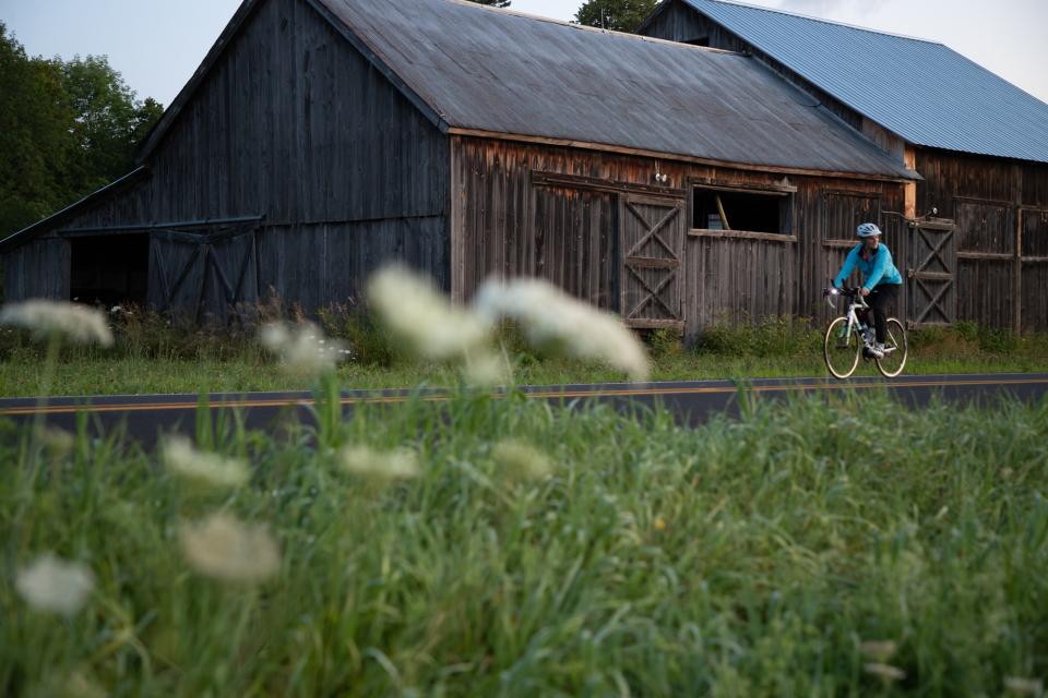 A woman rides a bicycle on a road past a historic barn in the Lake Champlain Region