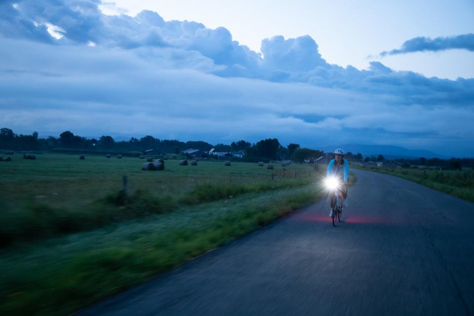 A woman rides a bicycle on a road at twilight with the bike's headlight switched on in the Lake Champlain Region