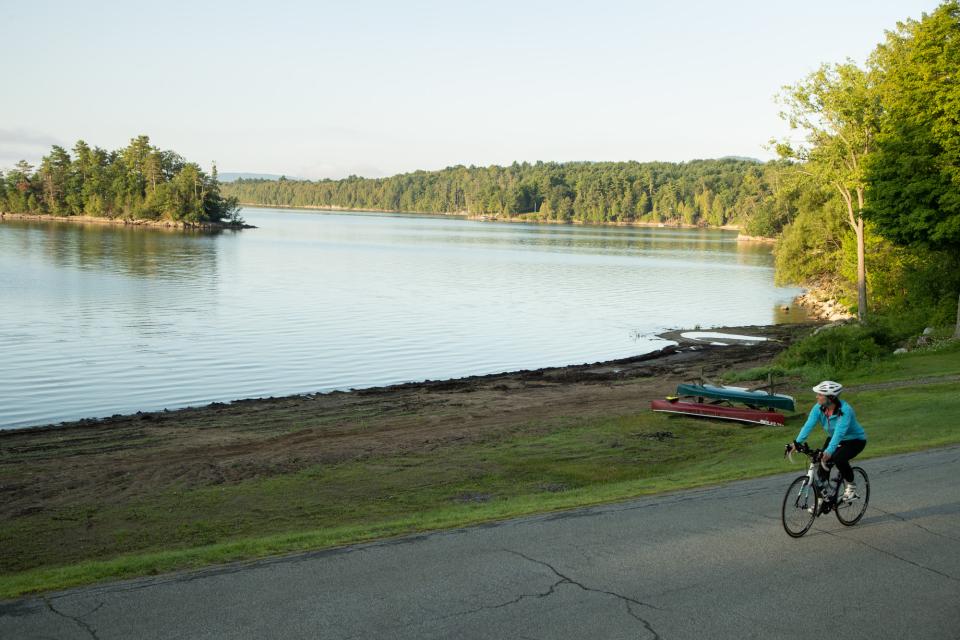 A woman rides a bicycle on a road past a body of water in the Lake Champlain Region