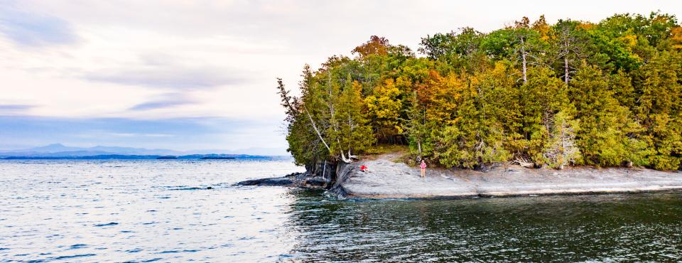 A peninsula in the Lake Champlain region, viewed from the water of Lake Champlain, with colorful foliage on land in the distance