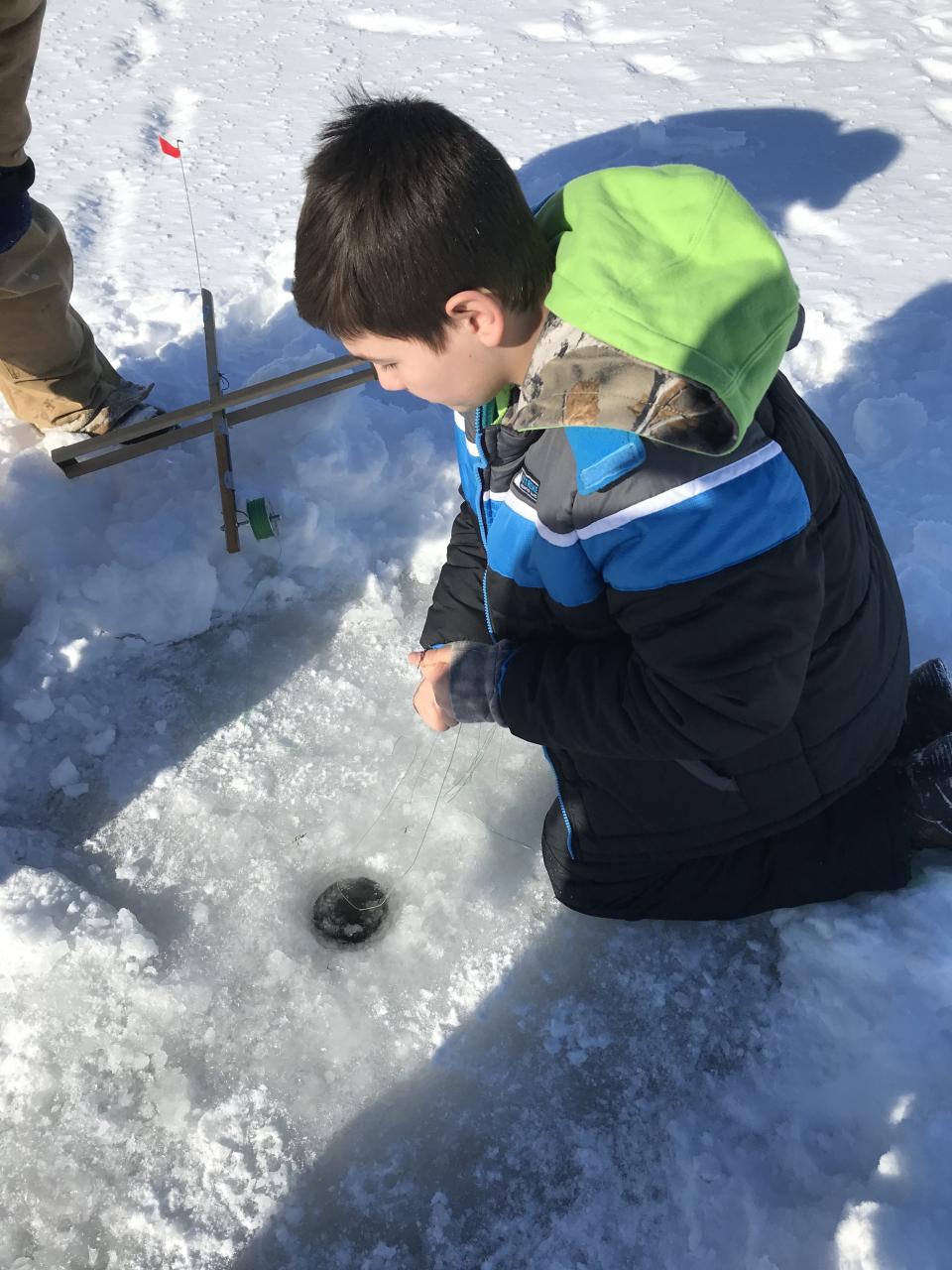 image of a young boy in winter wear looking into a drilled hole in the ice with other people around him