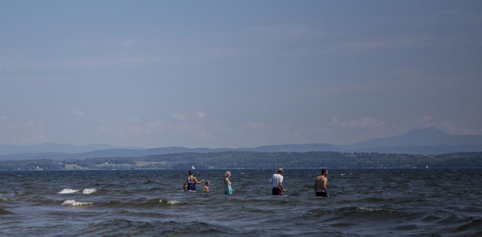 Adults and children play in the waters of Lake Champlain with the Green Mountains beyond.
