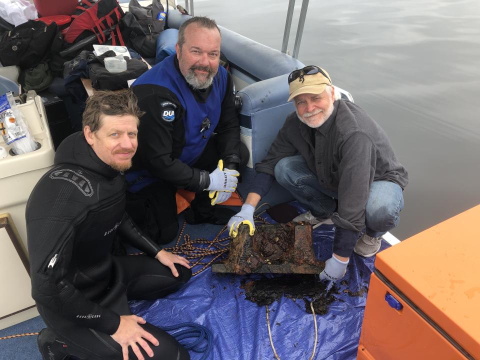 Two men in scuba gear and one other man pose on a boat with a rusty block of iron. Image courtesy Lake Champlain Maritime Museum.