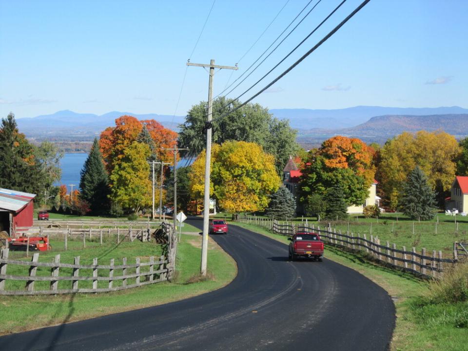 Red, orange, and yellow trees stand against a blue sky with a lake and mountains in the background