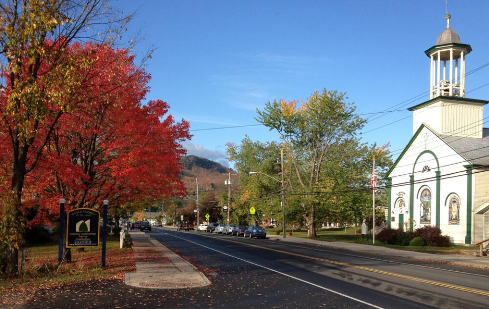 A tree with bright red leaves stands out along the side of a street. A tall white building is across the road.