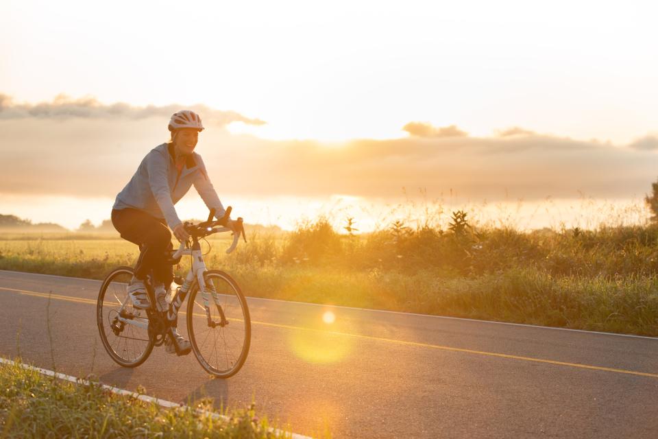 A woman rides a bike on the road as the sun rises behind her