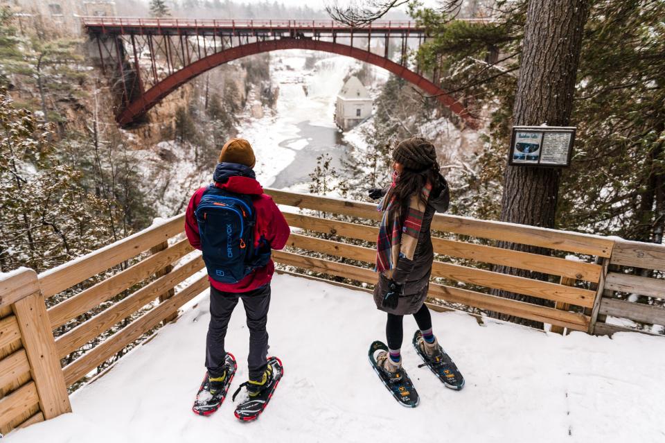 A man and woman in snowshoes and winter clothing look out at the bridge spanning Ausable Chasm