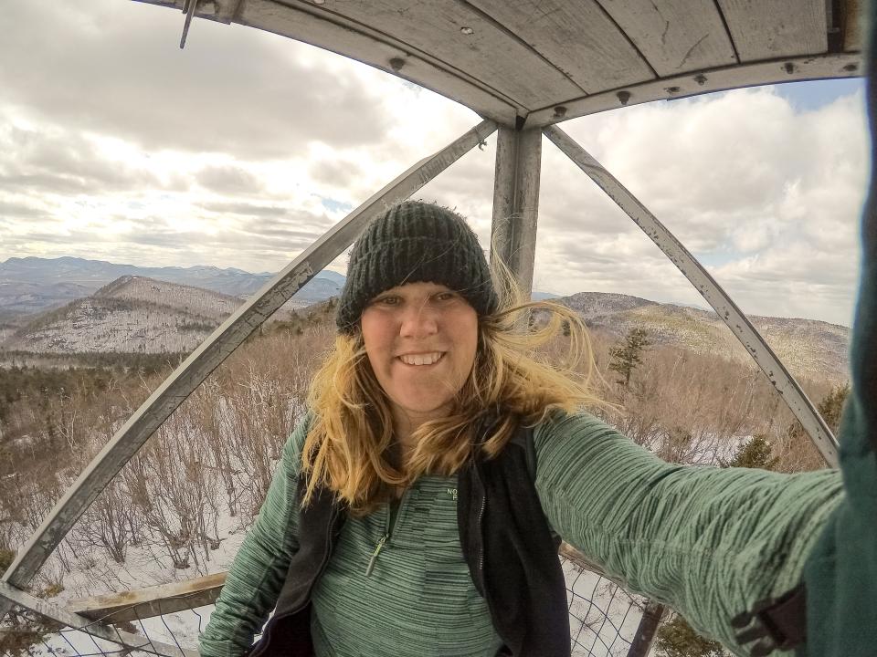 A self-portrait photo of a woman with long blonde hair in the cab of a fire tower on a mountain summit.