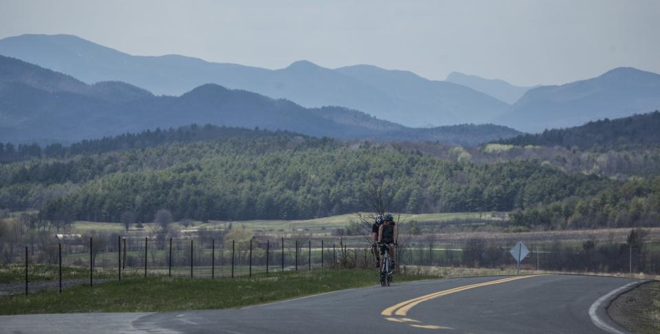 Two riders pedal up a rolling hill. The Adirondack High Peaks sit in the distance.