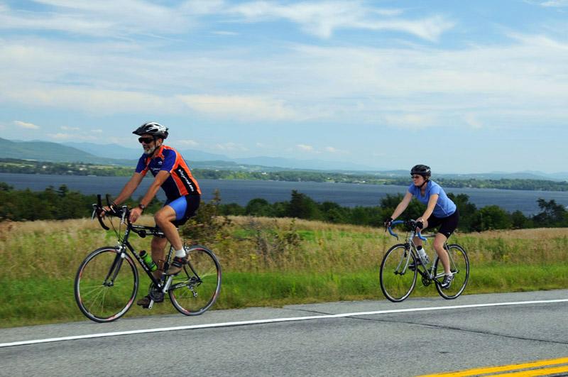 Two riders pedal along the road with water and the mountains looming behind them.