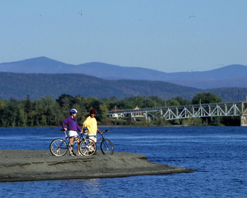 A man and woman stand next to their bikes on the shores of the lake, the mountains in the background.