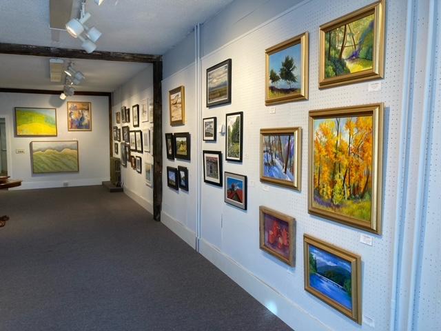 The interior of an art gallery room, with many paintings hanging on white-painted walls.