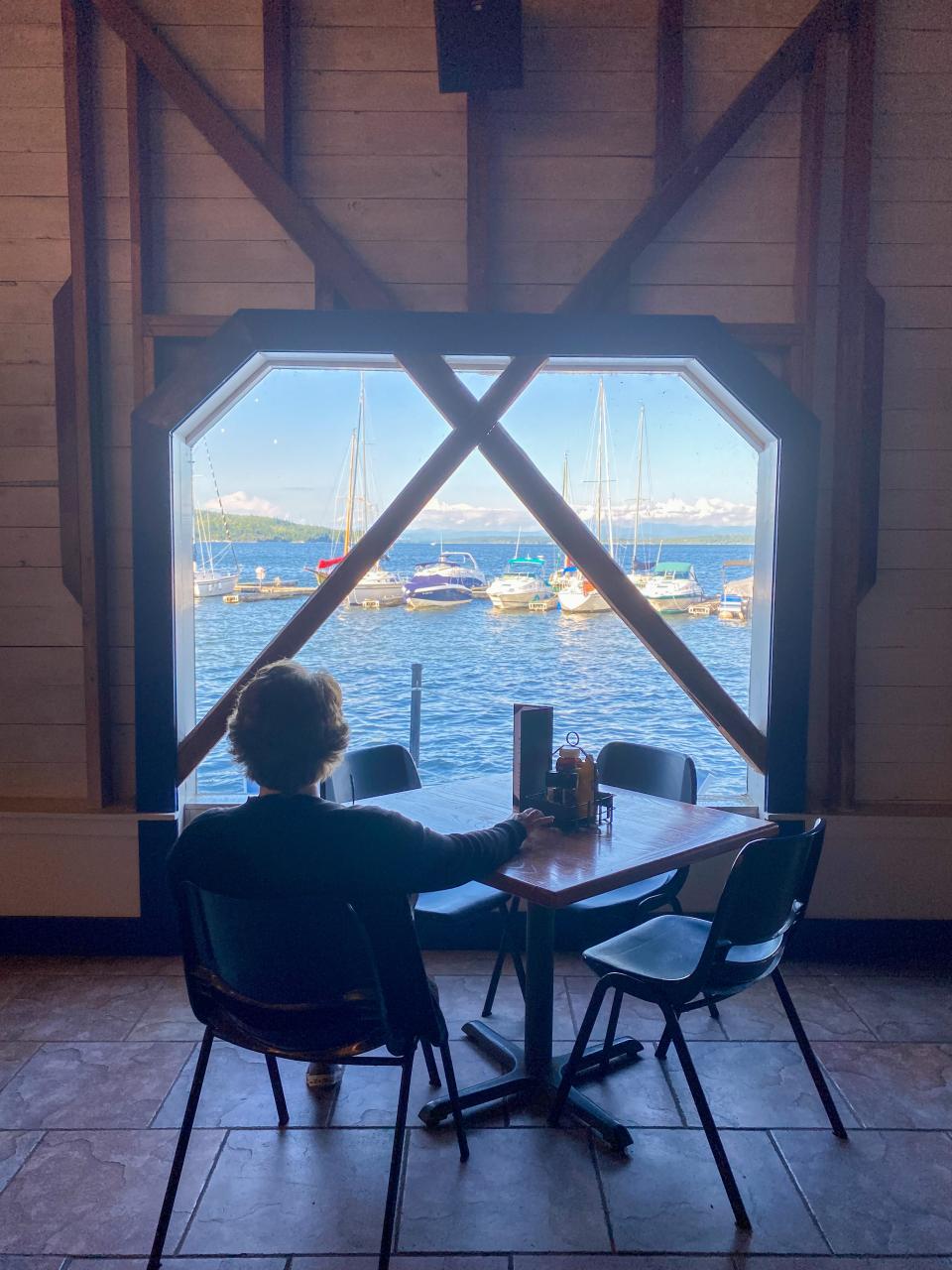 A woman at a restaurant table looks out a large window to a lake view.