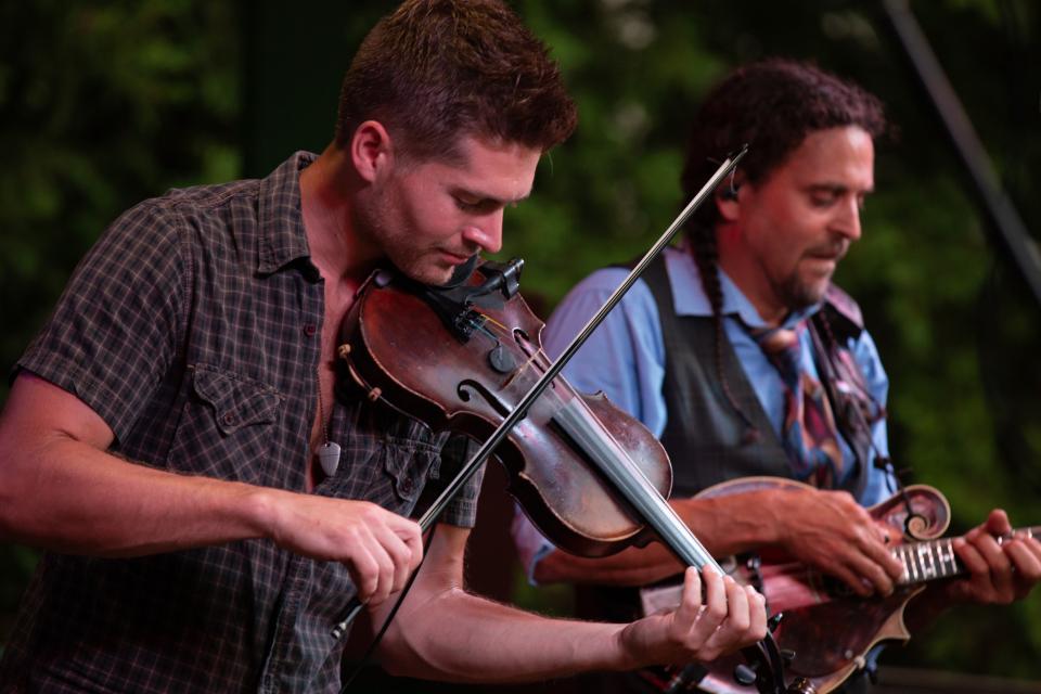 A man plays the fiddle during a live music performance. A mandolin player performs in the background.