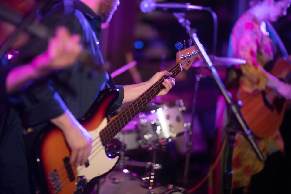 A closeup image of a person playing the electric guitar with drums in the background