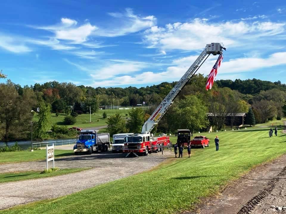 A large American flag hangs from the bucket of a fire truck at a park on a sunny day.