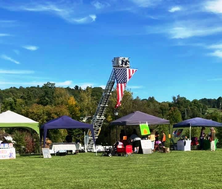 A fire truck and vendor tents at a grassy, tree-lined park on a sunny day.