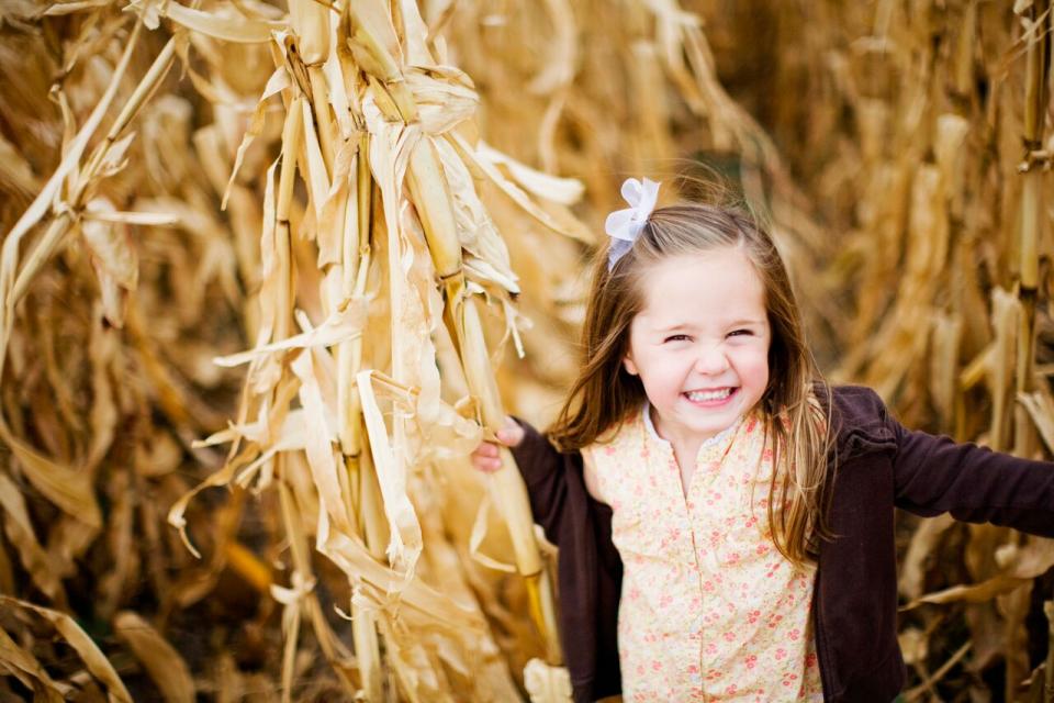A young girl stand smiling next to a brown cornstalk.