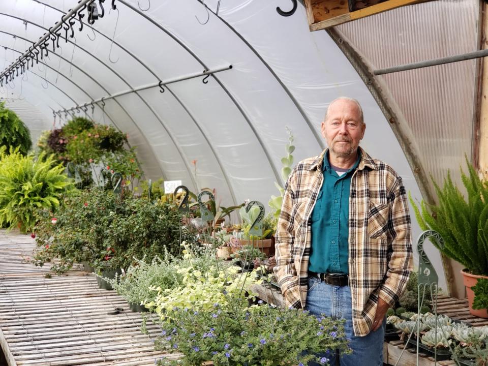 A man in a blue shirt and plaid jacket stands in a greenhouse next to plants.