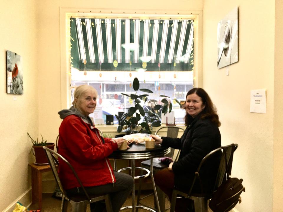 Two women sit for food and coffee at a window-side table.