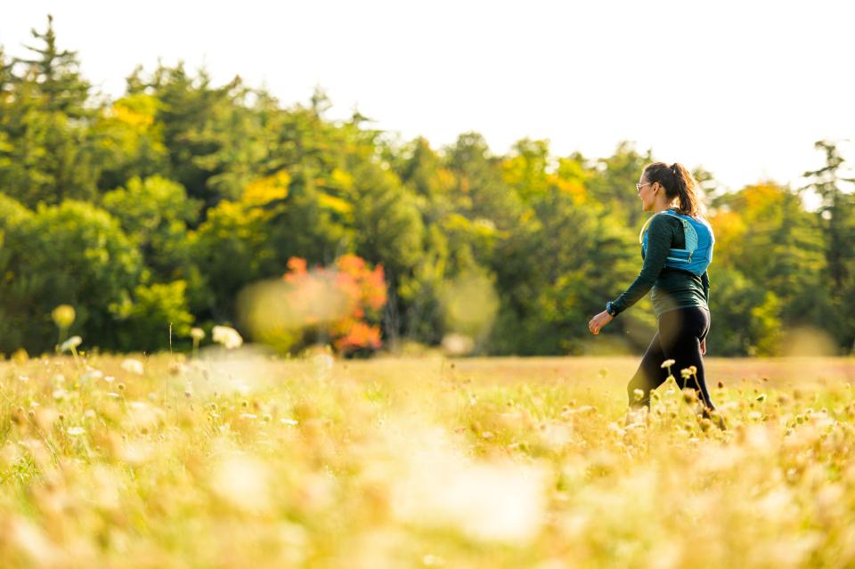 A tall woman in black clothing, wearing a bright blue backpack, walks across a golden-colored field in fall.