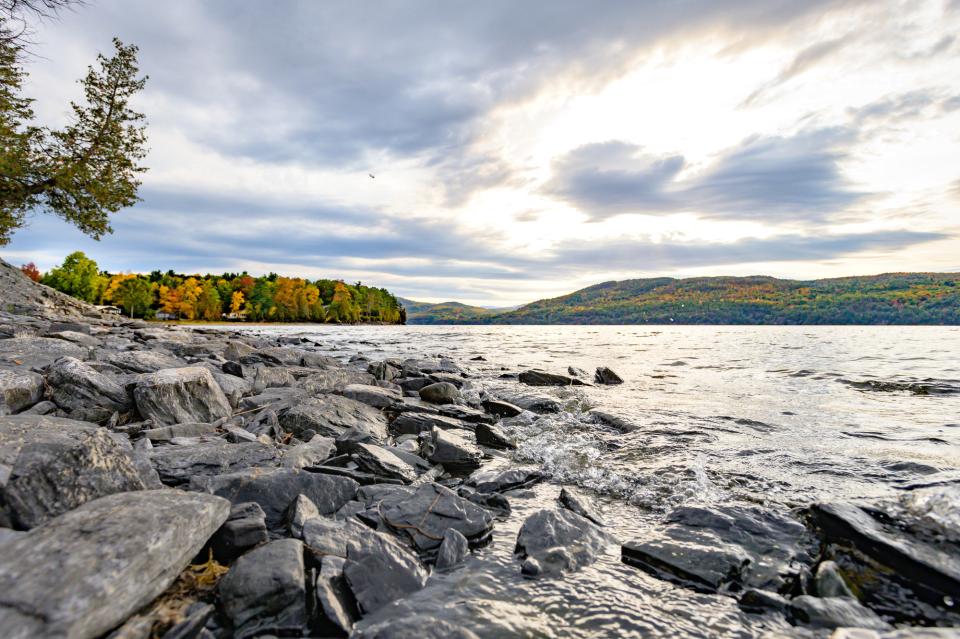 A view of Lake Champlain's rocky shoreline on a chilly autumn day, with some fall foliage colors along the distant shore