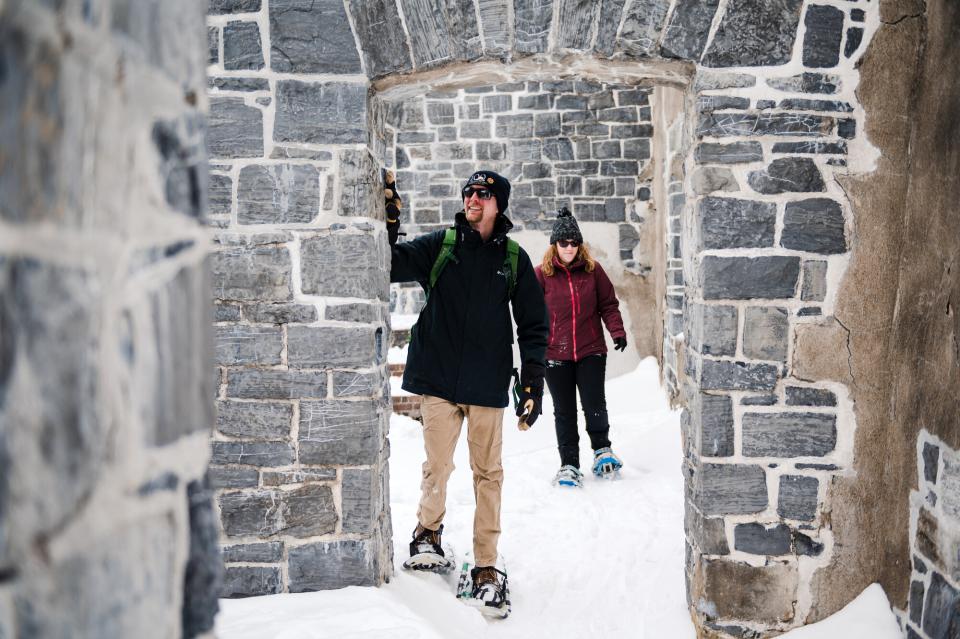 A man and woman in winter clothing snowshoe through the stone ruins of an 18th century fort.
