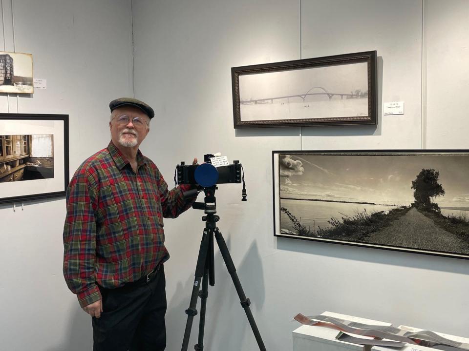 A man stands next to a camera on a tripod in front of a display of monochrome photos hung in a gallery at Ti Arts: Downtown Gallery