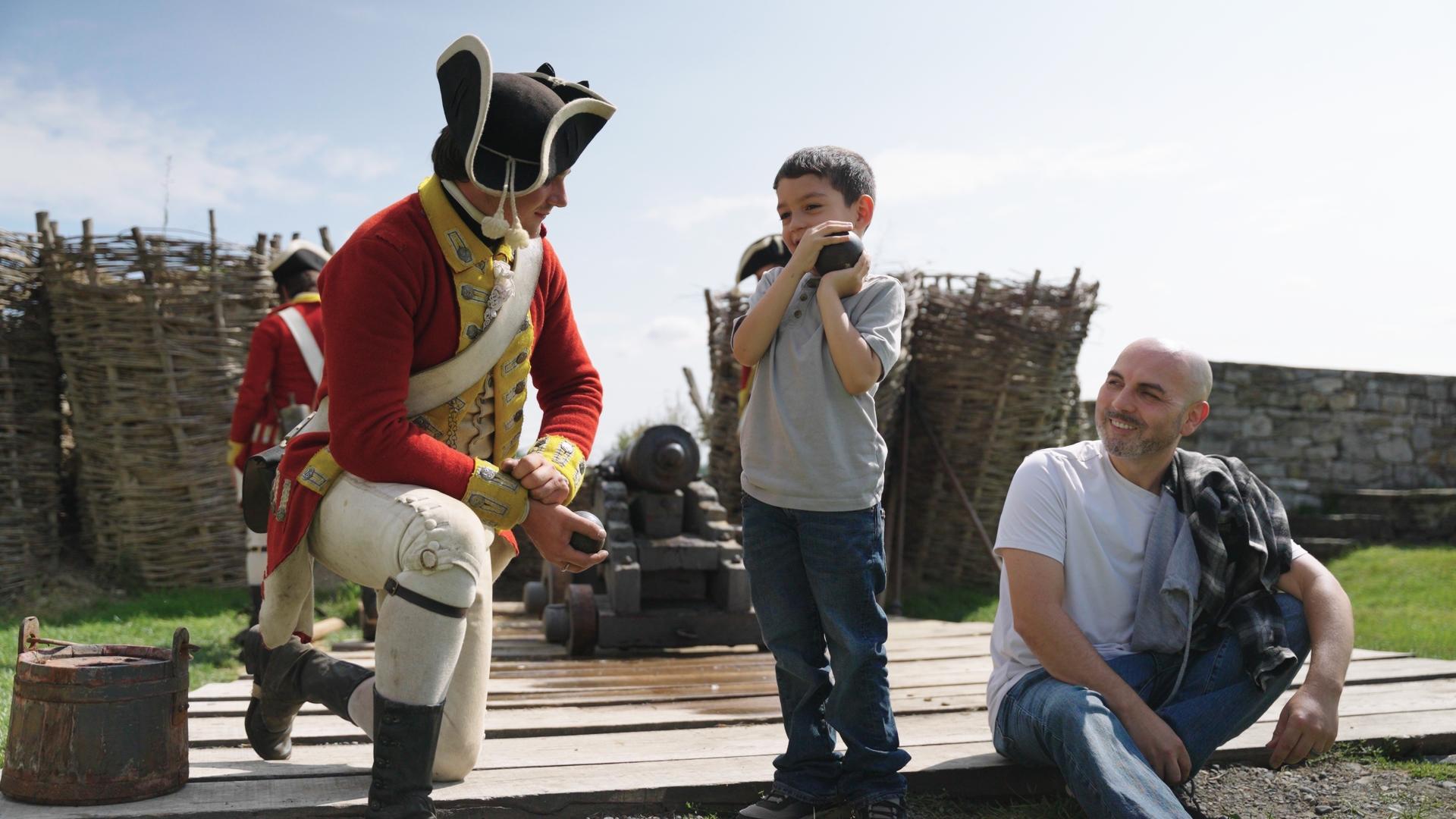 A reenactment performer in redcoat attire speaks with a young child.