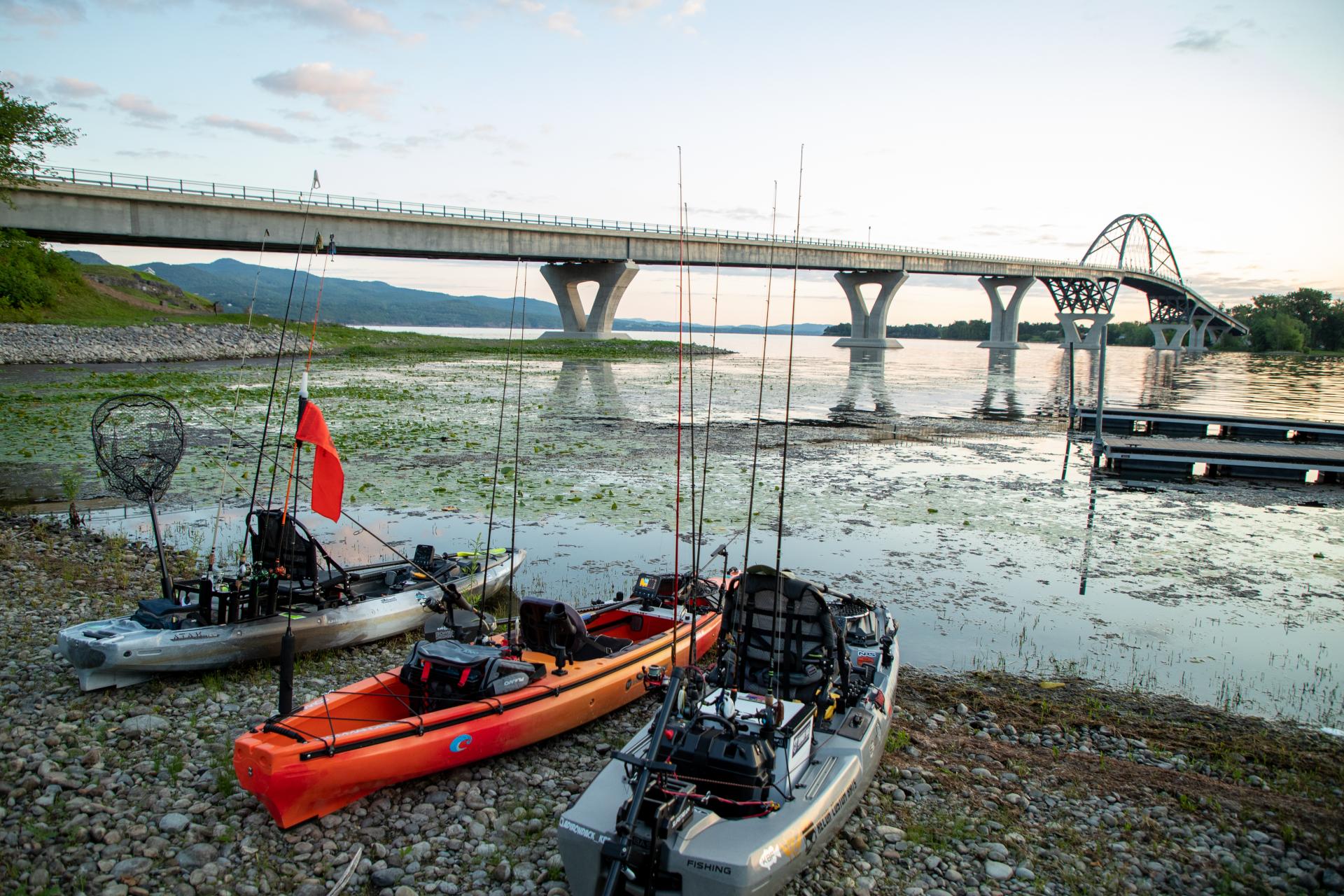 Kayaks fitted with trolling rods lined up on the edge of the water by a bridge.